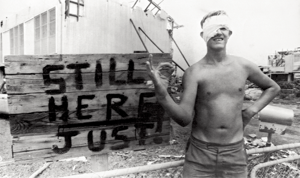 Photograph of a young man with a bandaged head making the ‘ok’ sign in front of destroyed buildings and a handpainted sign that reads ‘Still here, just!’