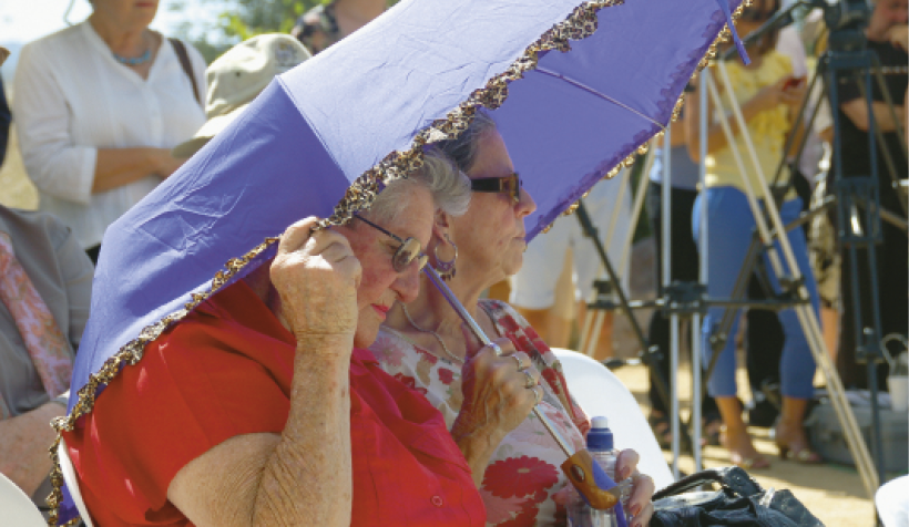Two elderly women are shading themselves with an unbrella. There are more people standing in the background.