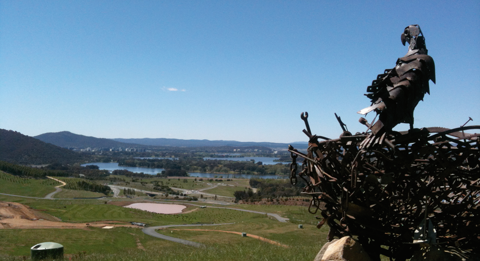 In the foreground is a metal sculpture of an eagle standing on a nest. In the background is the arboretum and the city of Canberra.