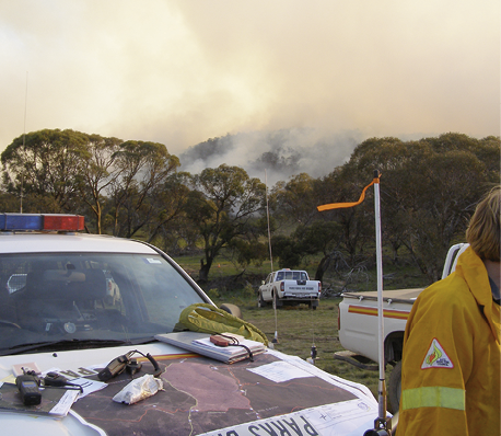 An emergency services vehicle in the foreground with equipment and maps on the bonnet. In the background is smoke from a controlled fire on a hillside.