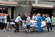 A photo of the front of the Christchurch Hospital Emergency Department. Several people are standing or walking in front, and hospital personnel are moving a patient on a stretcher.
