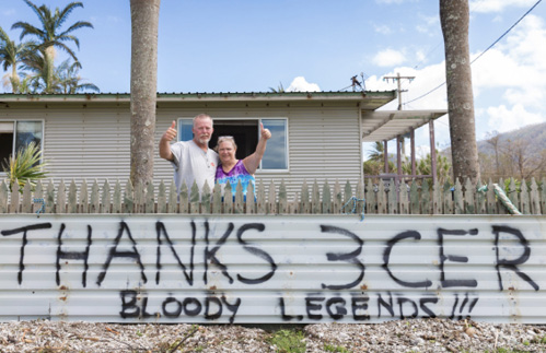 Image of two people standing in front of a home and behind a fence, holding their thumbs up. On the fence are the words ‘Thanks 3CER. Bloody legends’.