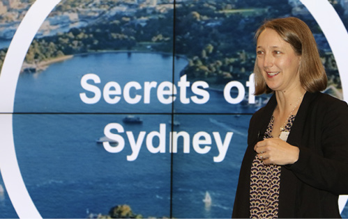 Image of Beck Dawson speaking in front of an aerial photo of Sydney, with the words ‘Secrets of Sydney’ on it.