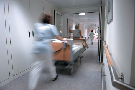 Two hospital staff are wheeling a hospital bed along a corridor.