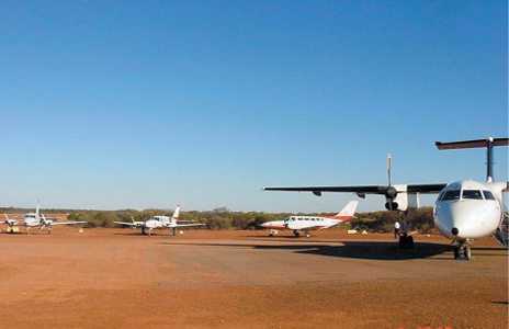 A red earth tarmac where four small propellor-driven aircraft are parked.