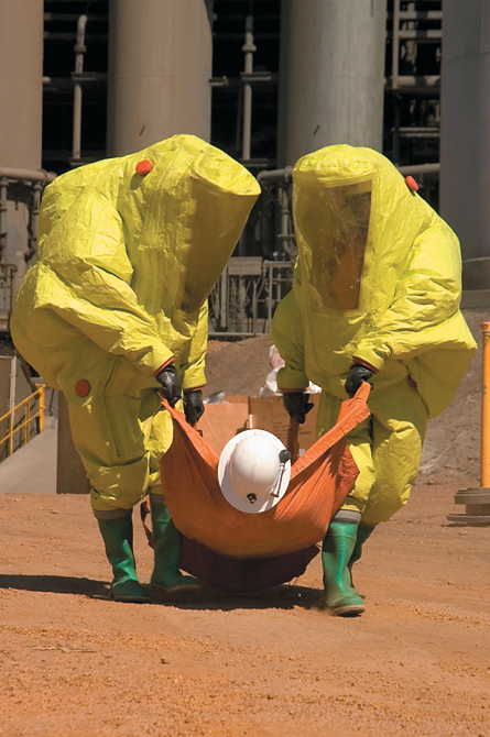 Two people dressed in yellow Hazmat coveralls, rubber boots and gloves are carrying a person on a tarpaulin away from an industrial facility.