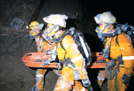 Four people wearing yellow coveralls, headlamp helmets and breathing aparatus are carrying an empty stretcher inside a cave.