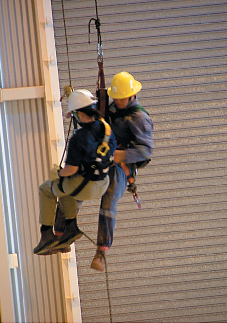 A man and a woman wearing work clothes and hard hats are suspended by ropes and harnesses next to a commercial building wall.