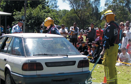 Two firemen demonstrating putting out a fire in a white sedan car with a crowd of people watching in the background