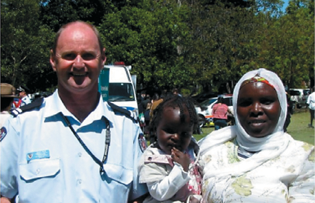 A male emergency services officer wearing a blue uniform smiling with an African woman and her toddler daughter