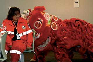 A red chinese pantomime dragon is headbutting a woman wearing an orange emergency services uniform 
