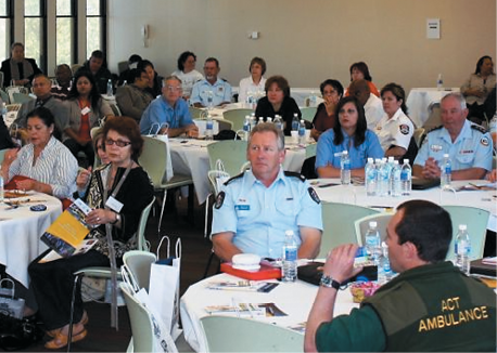 A large group of adults wearing emergency services uniforms or civilian clothing seated at round tables in a large conference room.
