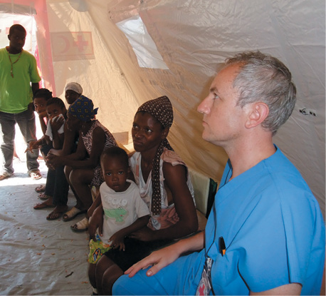 A causasian man in blue scrubs uniform is sitting with a small group of Haitian people inside a tent