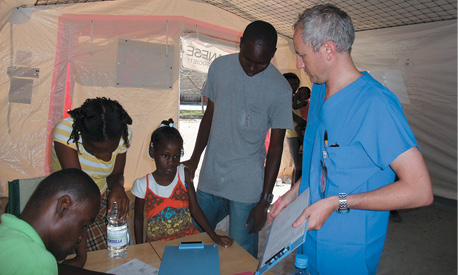 A causasian man in blue scrubs uniform is talking to a small group of Haitian people inside a tent