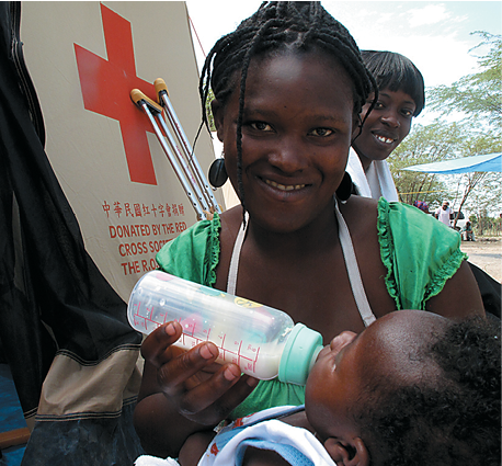 A Haitian woman feeding her baby a bottle of milk in front of a red cross sign.