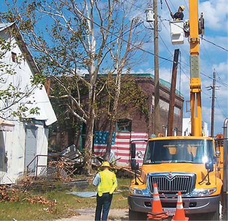 A truck with a man in a cherry-picker working on telegraph wires. In the background is a house and a warehouse building with a US flag mural.
