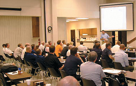 2 dozen adults, mostly men, seated in a large room watching a presentation.