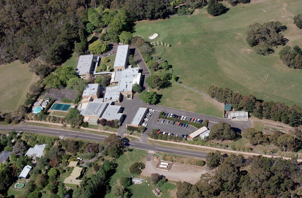 An aerial view of a collection of buildings, carparks, tanks, sheds, tennis court and other facilities surrounded by gardens, golf course and bush.