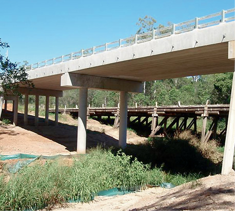 A new concrete bridge stands next to an old timber bridge over a weed-choked creek with forest in the background. There are some sediment barriers along the banks of the creek.