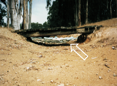 Side view of a dry creek bed spanned by a simple wooden bridge and beyond it a wall of sandbags. In the background are a stand of mature trees.