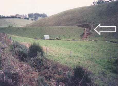 A grassy valley is spanned by a substantial earth dam wall. At one side of the dam wall there is significant erosion caused by overflow from the dam.