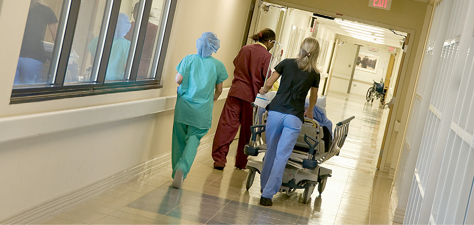 Three uniformed staff are wheeling a patient in a bed along a hospital corridor