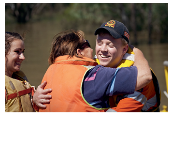 A smiling male emergency services officer hugs a woman while a smiling girls looks on. All are wearing marine life vests. In the background is brown floodwater and partially submerged bush.  