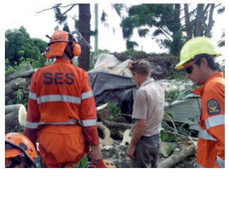 A middle-aged man and two emergency services workers contemplate sawn-up trees and wreckage.