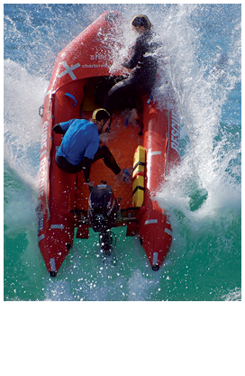 Two people are in a rigid hull inflatable outboard boat crashing almost vertically into a breaking wave.