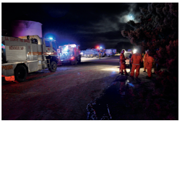 A night scene of several fire trucks with flood lights on, lined up along a dirt road. Several emergency services personnel are standing near some trees at the side of the road.
