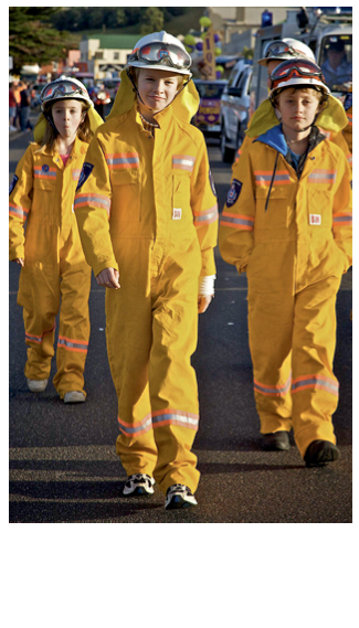 Four children in yellow coveralls, helmets and goggles are walking in a parade of vehicles along an urban street.