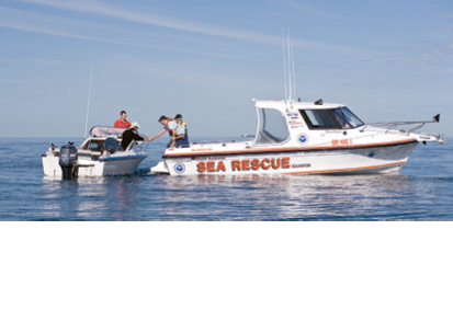 Two uniformed people on a sea rescue boat are assisting two people in a smaller outboard boat on calm open water.