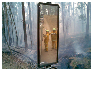 In the foreground, two firefighters are framed in the wing mirror of a firetruck. In the background is smouldering burnt forest.