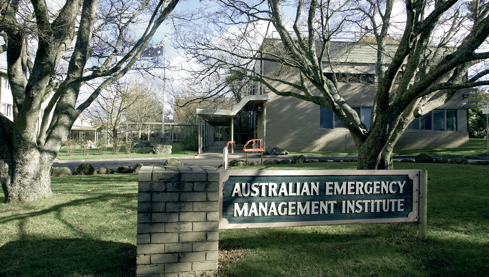 A two storey brick building with a grassed courtyard and Australian flag. In the foreground are two mature trees and the Australian Emergency Management Institute sign.