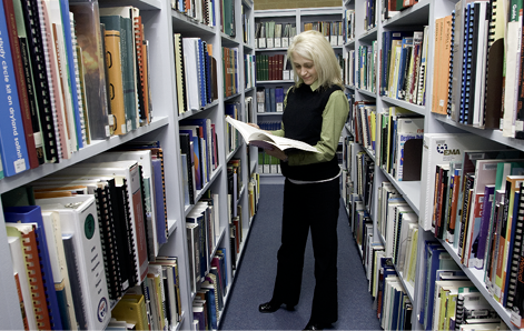 A woman reading a book in her hands is standing between rows of tall bookshelves.