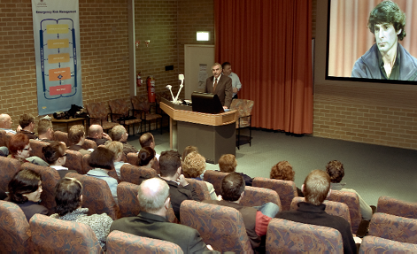 A man is speaking from a lectern in front of a large projection screen to an audience in tiered seating. 
