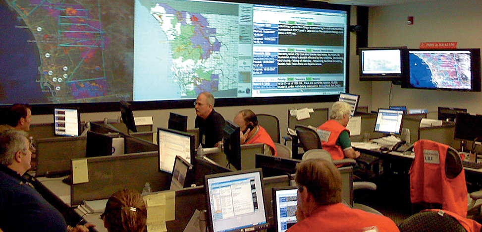 An incident response room - several people are seated at computer desks. In the background is a large screen showing several maps and information summaries.