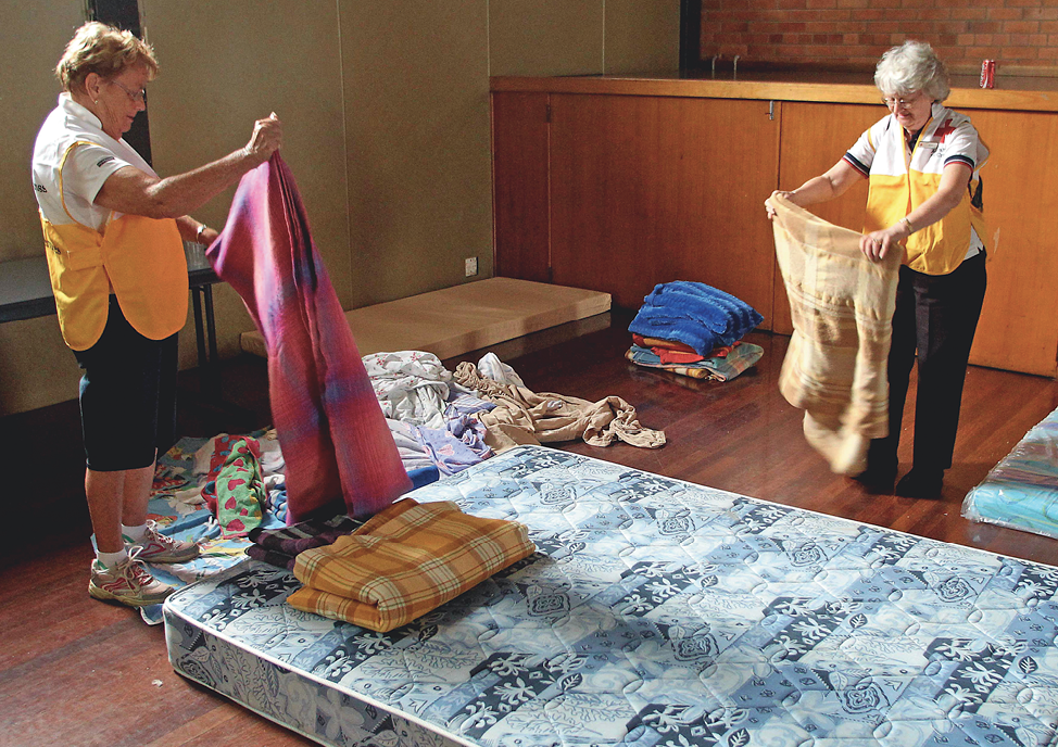 Two senior women wearing yellow vests are folding bed linen in a community hall. There are several mattresses on the floor.