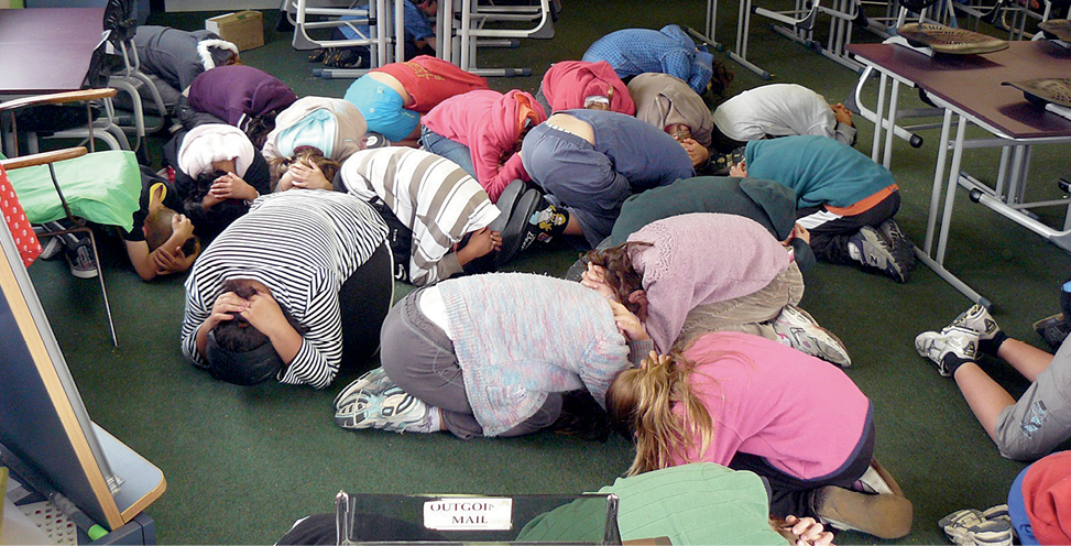 A group of children are crouched on the floor of a classroom, covering their heads with their hands.