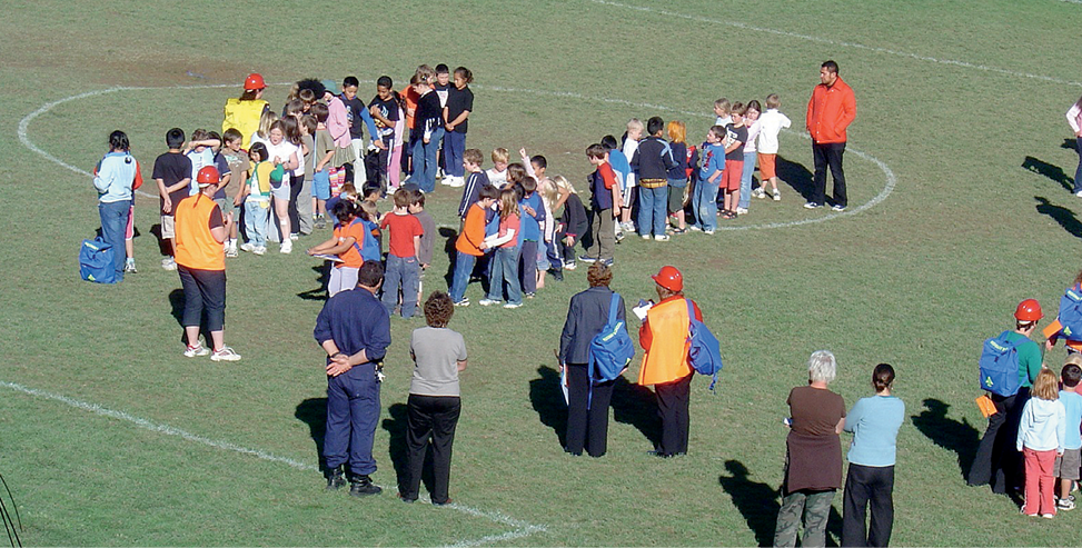 Groups of children are lined up on a grass playing field. Several adults are looking on. Some of the adults are wearing orange vests and red hardhats.