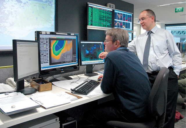 Photograph of two men looking at computer screens. The screens show coloured shapes representing a cyclone.