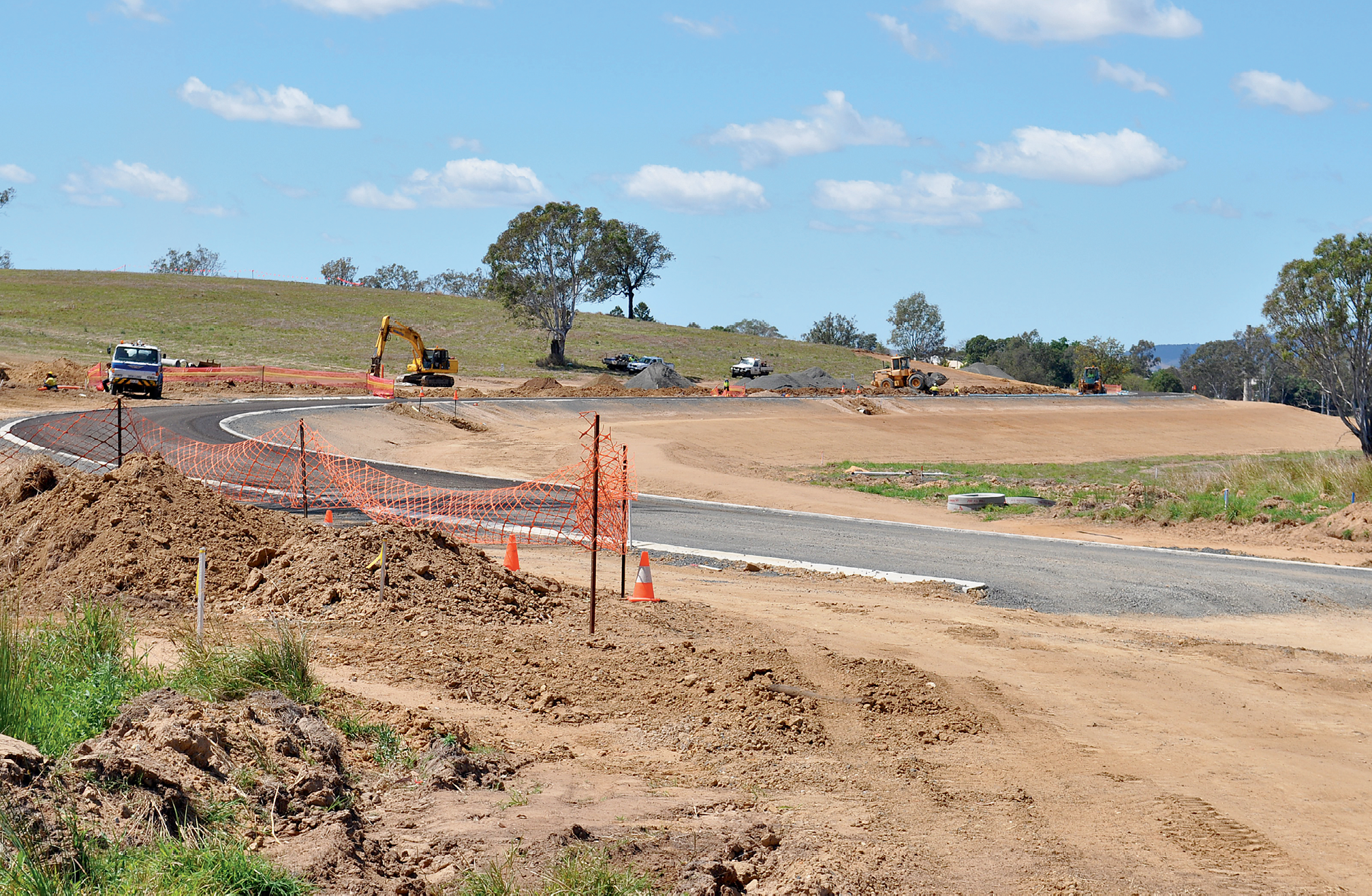 Photograph of road construction in the countryside. Piles of dirt are next to orange netting and traffic cones. A truck, digger and cars are in the background near a gum tree.