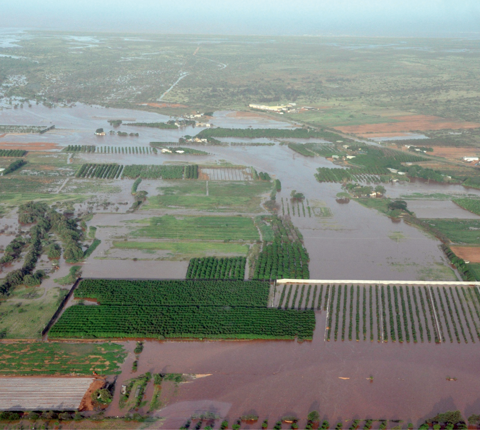 An aerial photograph of farms and buildings under floodwaters. The water is brown and the sky is blue. Some of the paddocks are underwater and the trees are poking up out of other areas.