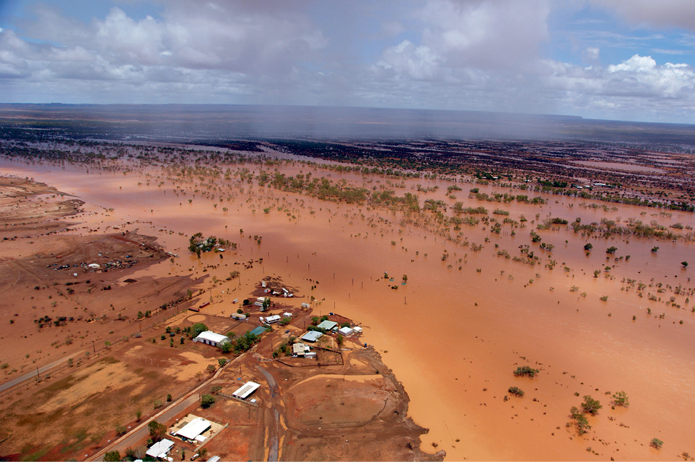 An aerial photograph of buildings and surrounding countryside under floodwaters. The water is brown and the sky is blue but cloudy.
