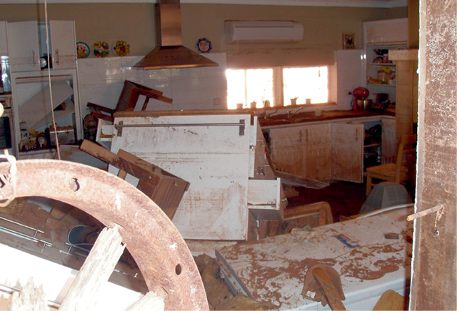 A photograph of the kitchen of a house that has been damaged by a flood. Some of the furniture is falling over and has detached from the wall. The white cupboards have been stained brown.