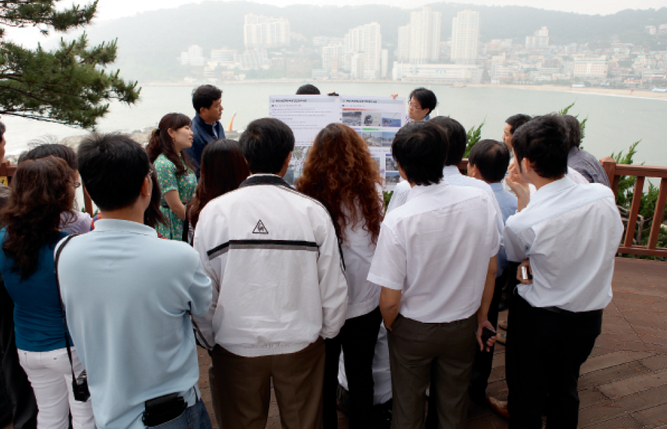 A photograph of a group of people from Vietnam looking at a poster being displayed by two people from the National Disaster Management Institute. The people are outside standing on a lookout with a city in the background