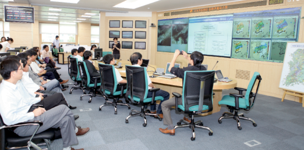 A photograph of people visiting the Meteorological Office in the Republic of Korea. About 15 people sit in chairs watching a woman give a PowerPoint presentation on a large screen. The screen shows weather patterns and cloud maps.