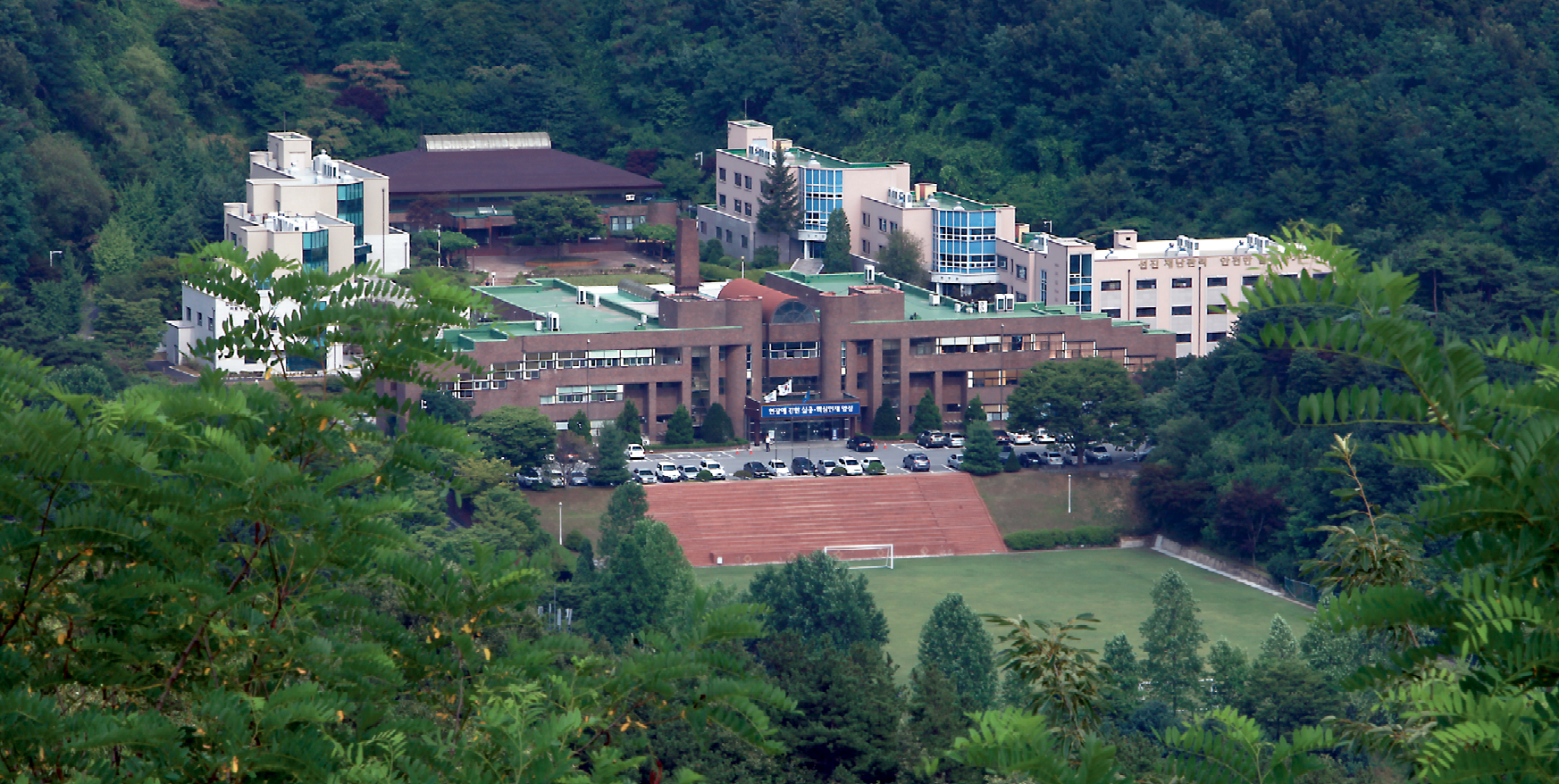 A photograph of the National Disaster Management Institute and National Fire Service Academy campus in the Republic of Korea. The buildings are brown and white, with steps leading down to a green sporting field. Lush green plants and trees surround the buildings.