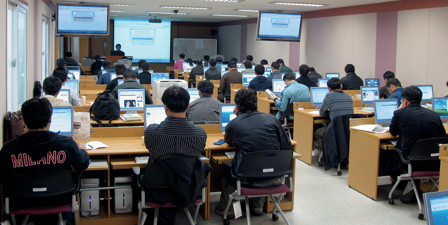 Students look at computers in a classroom at the National Disaster Management System Information Centre. A teacher at the front of the room is showing a PowerPoint presentation on a large screen.