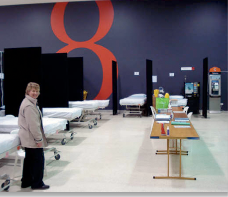 Photograph of hospital beds separated by free-standing screens in a large room. An administration table, payphone and vending machine are also in the room.